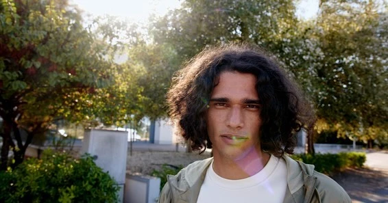 Portrait of a young adult male with curly hair outdoors during daytime with lush green trees and foliage in the background.