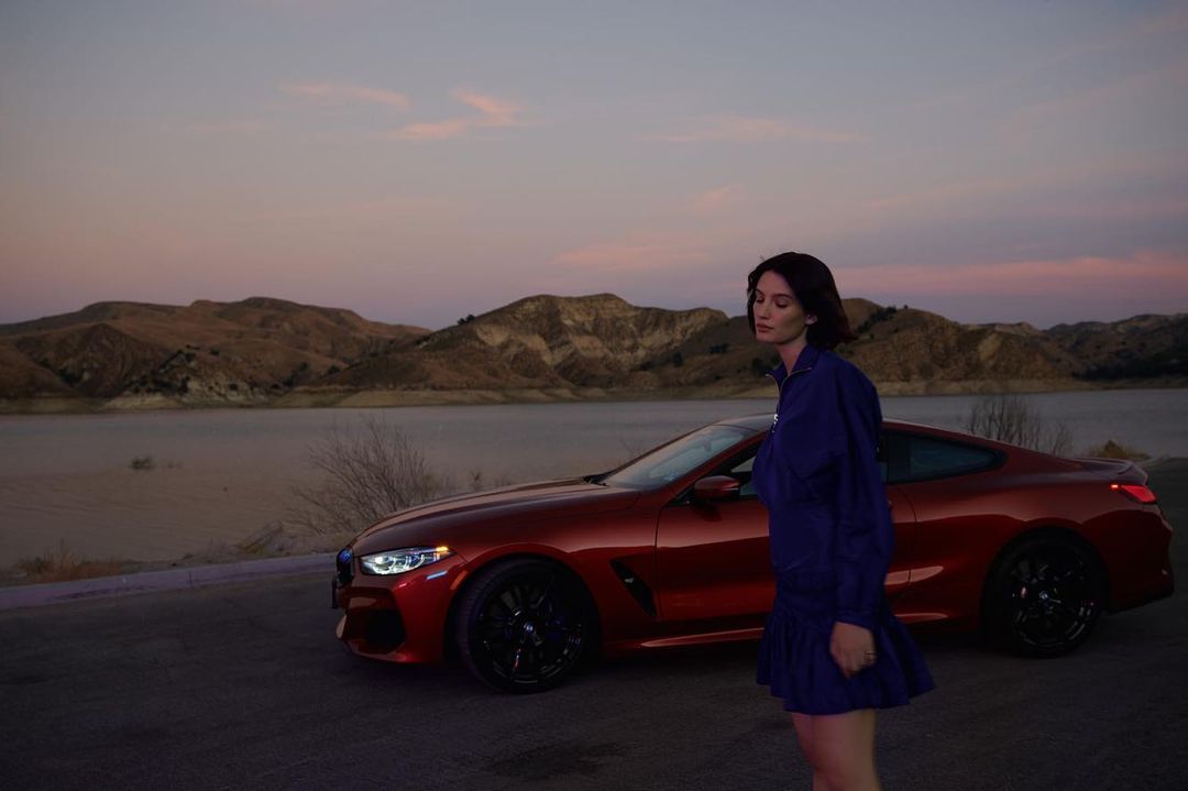 Woman standing outdoors next to a parked red sports car with hills and water under a twilight sky