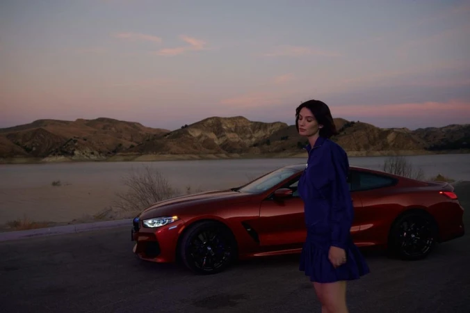 Woman standing outdoors next to a parked red sports car with hills and water under a twilight sky