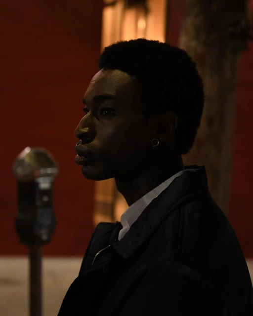 Portrait of a young Black man in a black jacket and white shirt against a red wall, captured in low lighting at eye level.