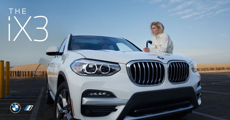 White BMW iX3 electric vehicle parked outdoors with an adult female standing next to it on a sunny day. Automotive marketing image.