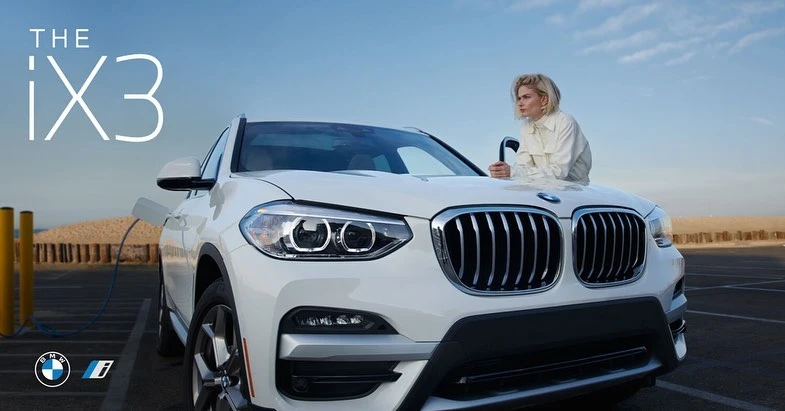 White BMW iX3 electric vehicle parked outdoors with an adult female standing next to it on a sunny day. Automotive marketing image.