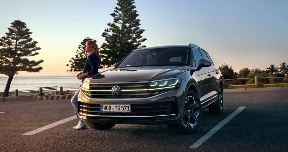 Dark grey Volkswagen Touareg SUV parked on asphalt next to adult female leaning on hood near coast, trees, clear sky at sunset.