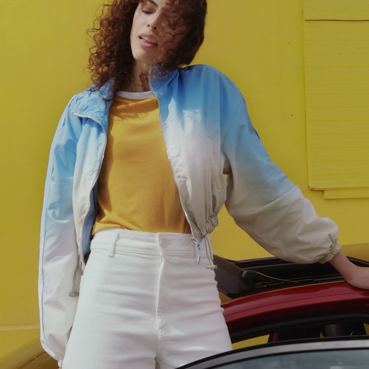 Stylish woman in cropped blue gradient jacket and white pants poses against a vivid yellow wall, showcasing a fashionable, casual look.