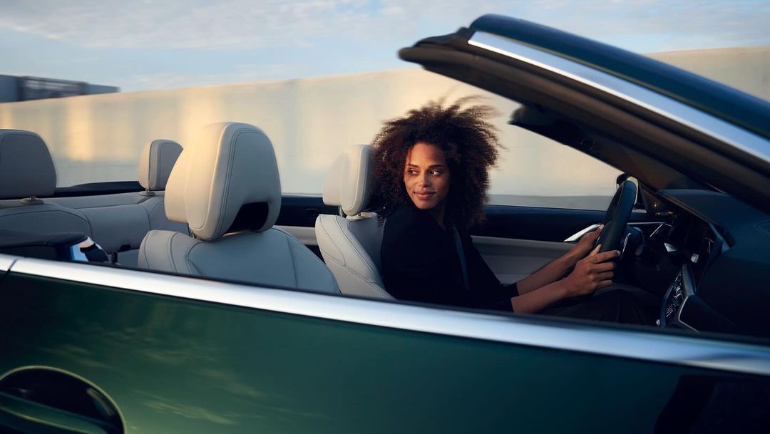 A woman with curly hair smiles and drives a convertible car along a highway in a bright, daytime setting with a blurred background.