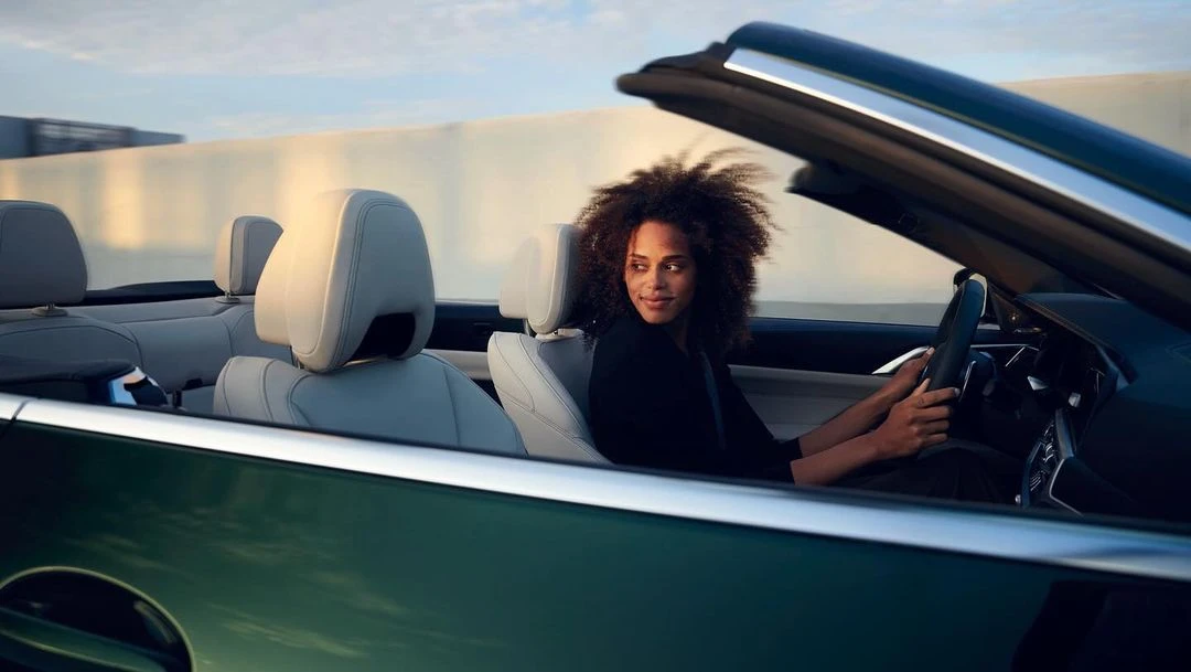 A woman with curly hair smiles and drives a convertible car along a highway in a bright, daytime setting with a blurred background.