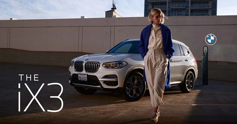 White BMW iX3 electric SUV car stands parked with a blonde adult woman in a blue coat in a parking lot during the daytime.