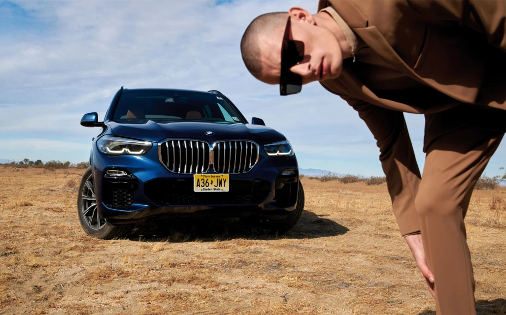 A deep blue BMW X5 SUV is parked on brown dry grass with a man in a brown suit and sunglasses leaning into the shot.