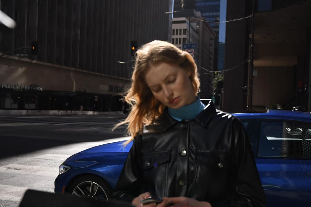 Young woman in a leather jacket checks her cell phone, standing on a sunny city street by a blue car, framed by modern buildings.