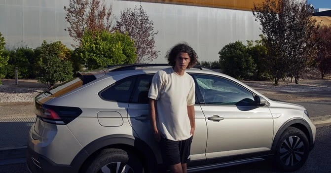 Young adult man stands next to a silver compact SUV in an outdoor setting with green trees and a building in the background.