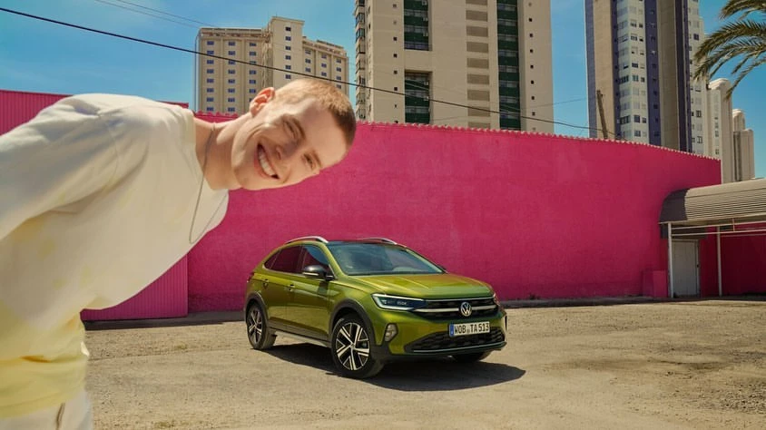 Smiling man looks at a green Volkswagen Taigo SUV in an outdoor setting against a pink wall and tall buildings under a clear blue sky.