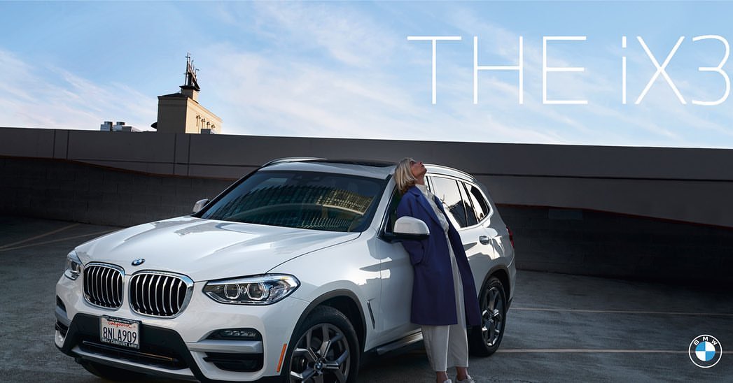 A white BMW iX3 is parked on a rooftop with a woman leaning against it under a blue sky, creating a scene that blends luxury and urban style.