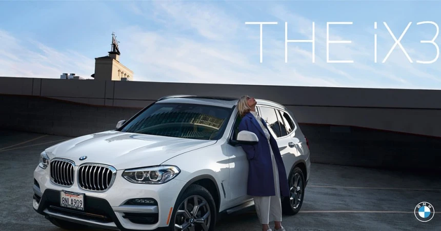 A white BMW iX3 is parked on a rooftop with a woman leaning against it under a blue sky, creating a scene that blends luxury and urban style.