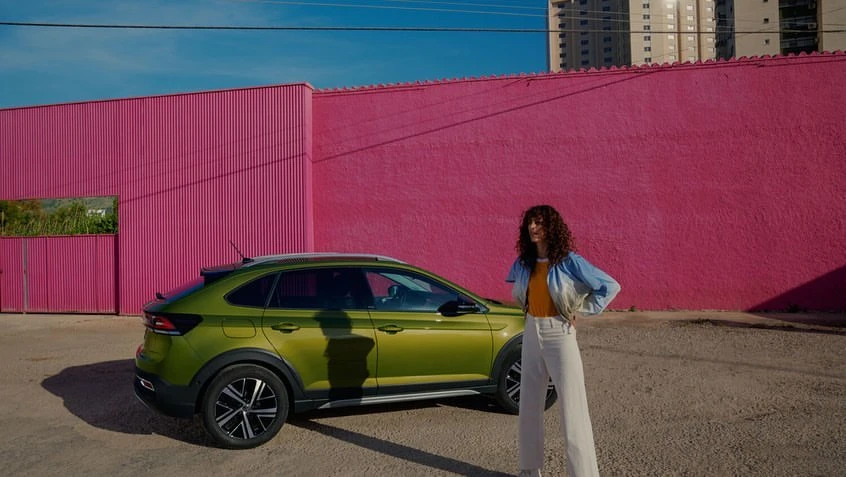 A green Volkswagen Taigo parked in front of a pink building with a brunette-haired woman standing beside it with an urban backdrop.