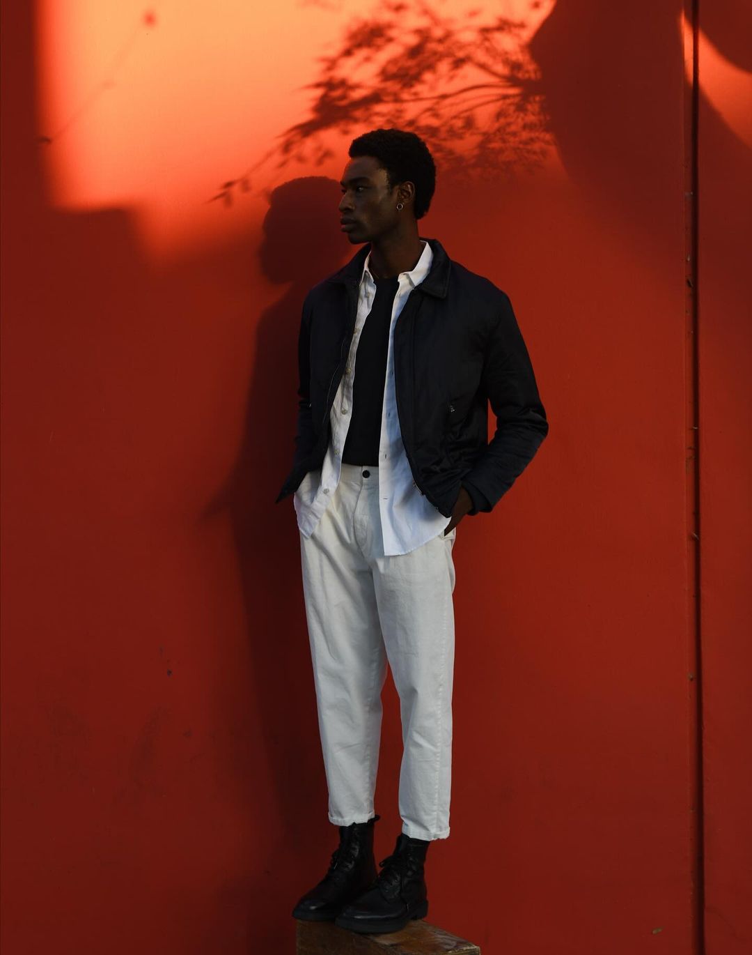 A stylish Black man stands against an orange wall in an open white shirt, dark jacket, and white pants, posing in dark boots.