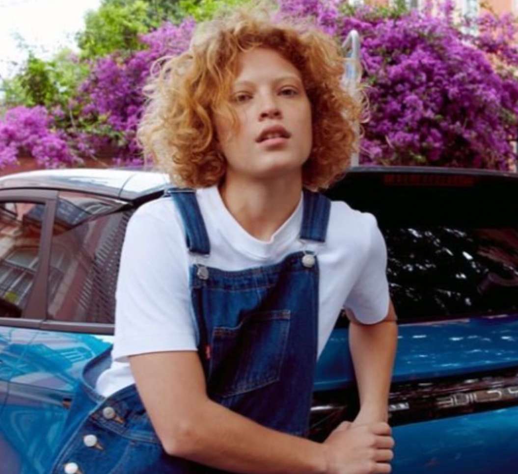 Young adult woman with curly hair poses by a blue car with purple flowers in the background for a fashion or beauty-themed lifestyle shot.