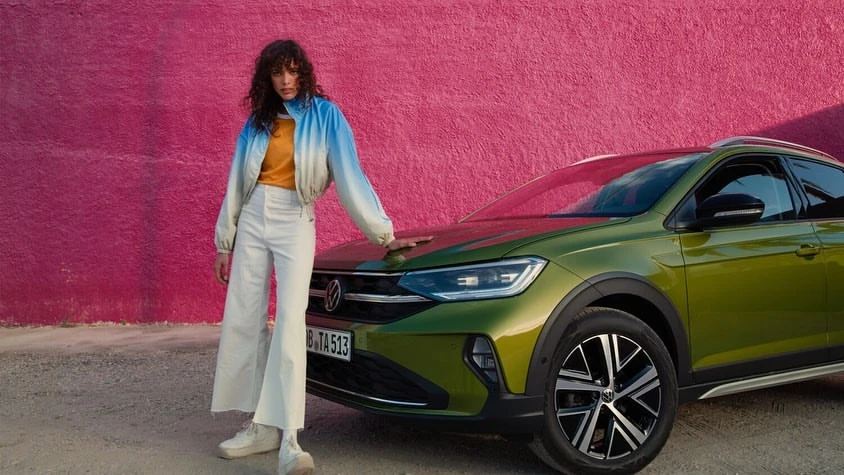 A woman in a jacket and wide-leg pants poses on the hood of a green Volkswagen car in front of a pink textured wall.