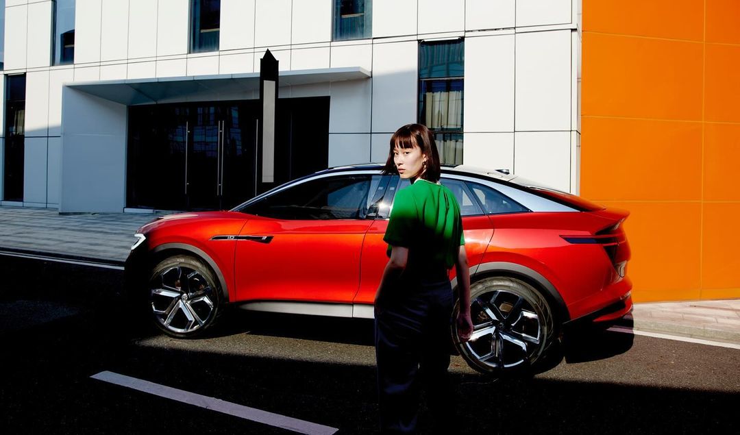 An adult female in a green shirt stands next to a red SUV in an urban environment with a modern building and orange wall backdrop.