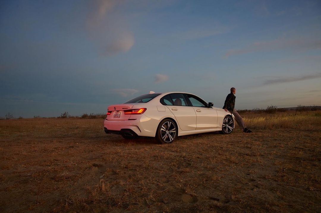 White BMW sedan parked in a field at dusk, with an adult male resting against the hood, under a sky with scattered clouds.