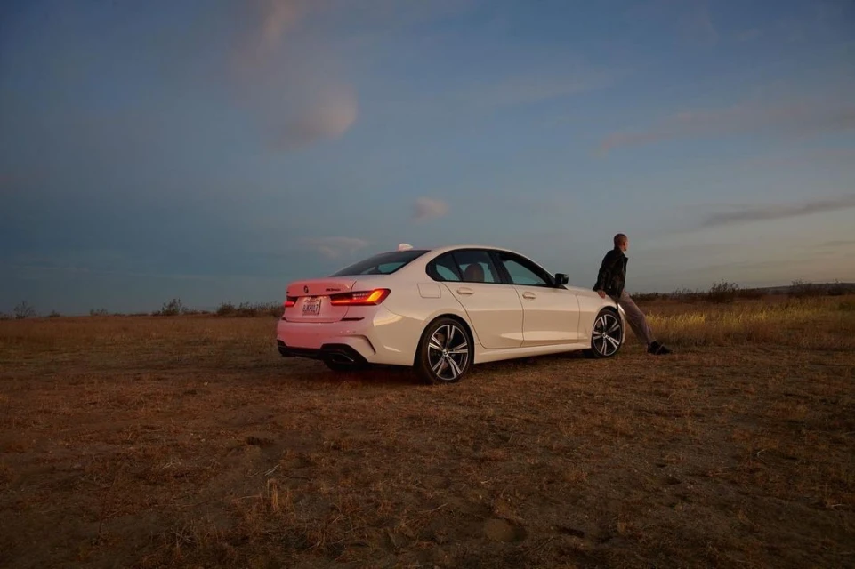 White BMW sedan parked in a field at dusk, with an adult male resting against the hood, under a sky with scattered clouds.