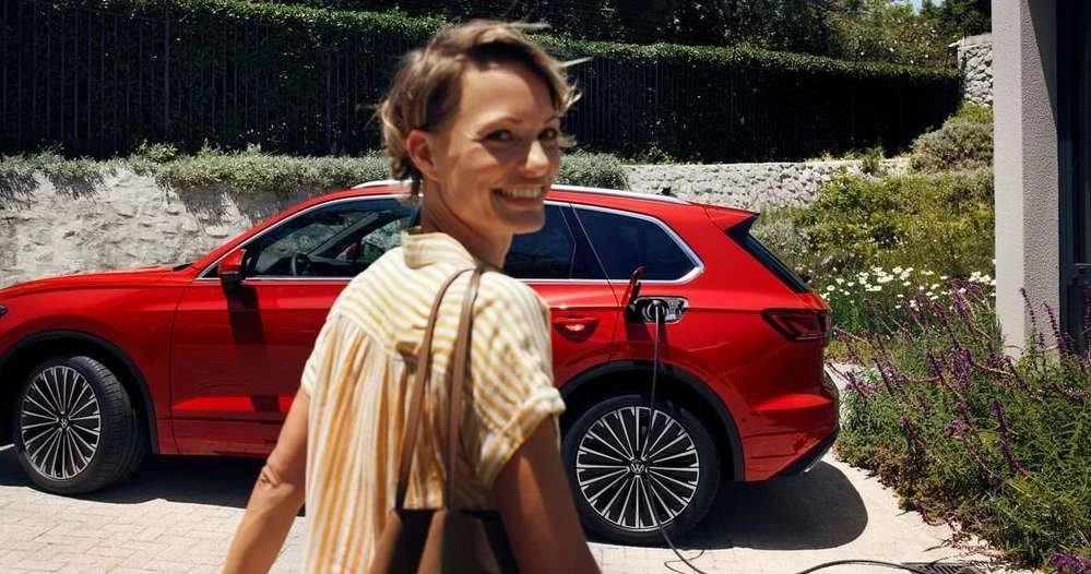 Woman smiles near a red electric SUV plugged into a house charger, showcasing eco-friendly automotive technology in a bright outdoor setting.