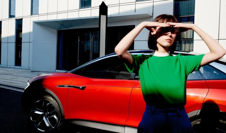 Adult female in a green top poses in front of a red electric car and a modern white building on a sunny day.