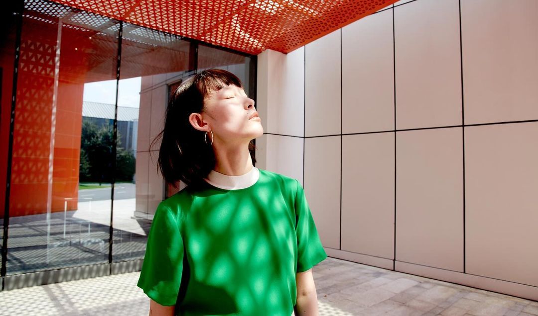 Woman in green top stands outdoors with sunlight casting patterned shadows, framed by modern architecture with orange and white panels.
