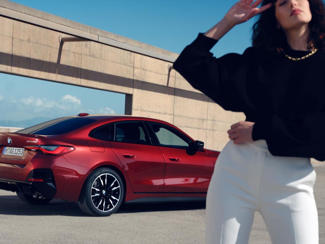 A stylish woman in white pants strikes a pose next to a sleek red BMW 4 Series Gran Coupe against a minimalist concrete backdrop.