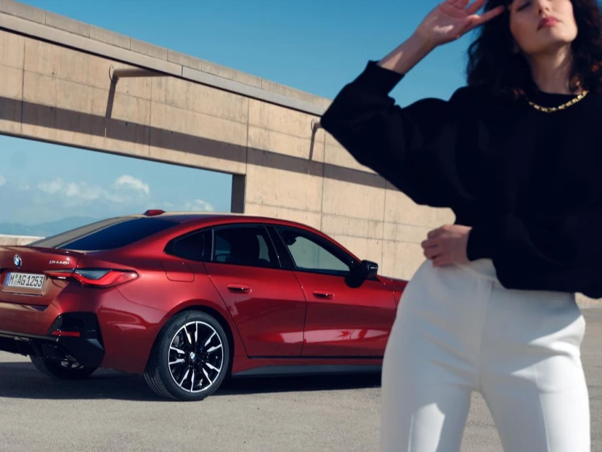 A stylish woman in white pants strikes a pose next to a sleek red BMW 4 Series Gran Coupe against a minimalist concrete backdrop.