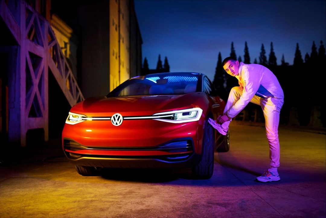 Red Volkswagen electric car with a man tying his shoe in an urban setting, lit with dramatic neon lights against a dark background.