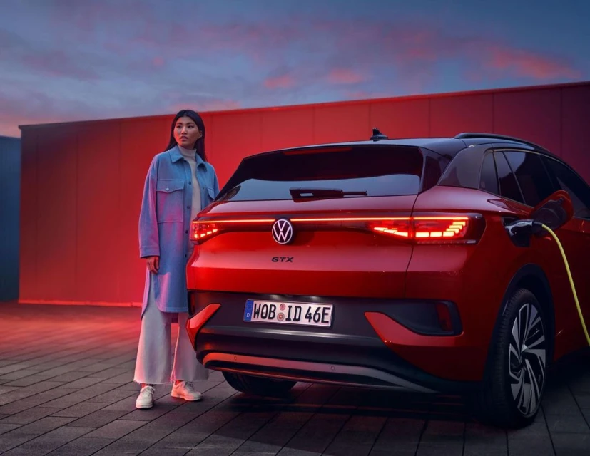 Rear view of a red Volkswagen ID.4 GTX electric car with an Asian woman standing alongside it, plugged into a charging station.