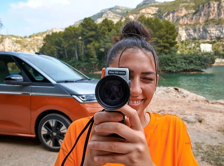 A woman in an orange shirt smiles while holding an analog camera up to her eye, with a van and mountains in the background.