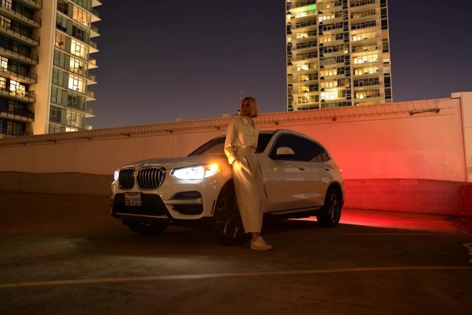 An adult female poses with a white BMW SUV in an urban environment at night with lit-up buildings in the background.