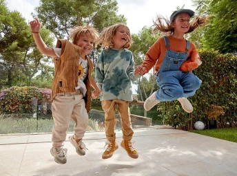 Three young children jumping in the air outdoors, holding hands and laughing with sunlight in their hair.