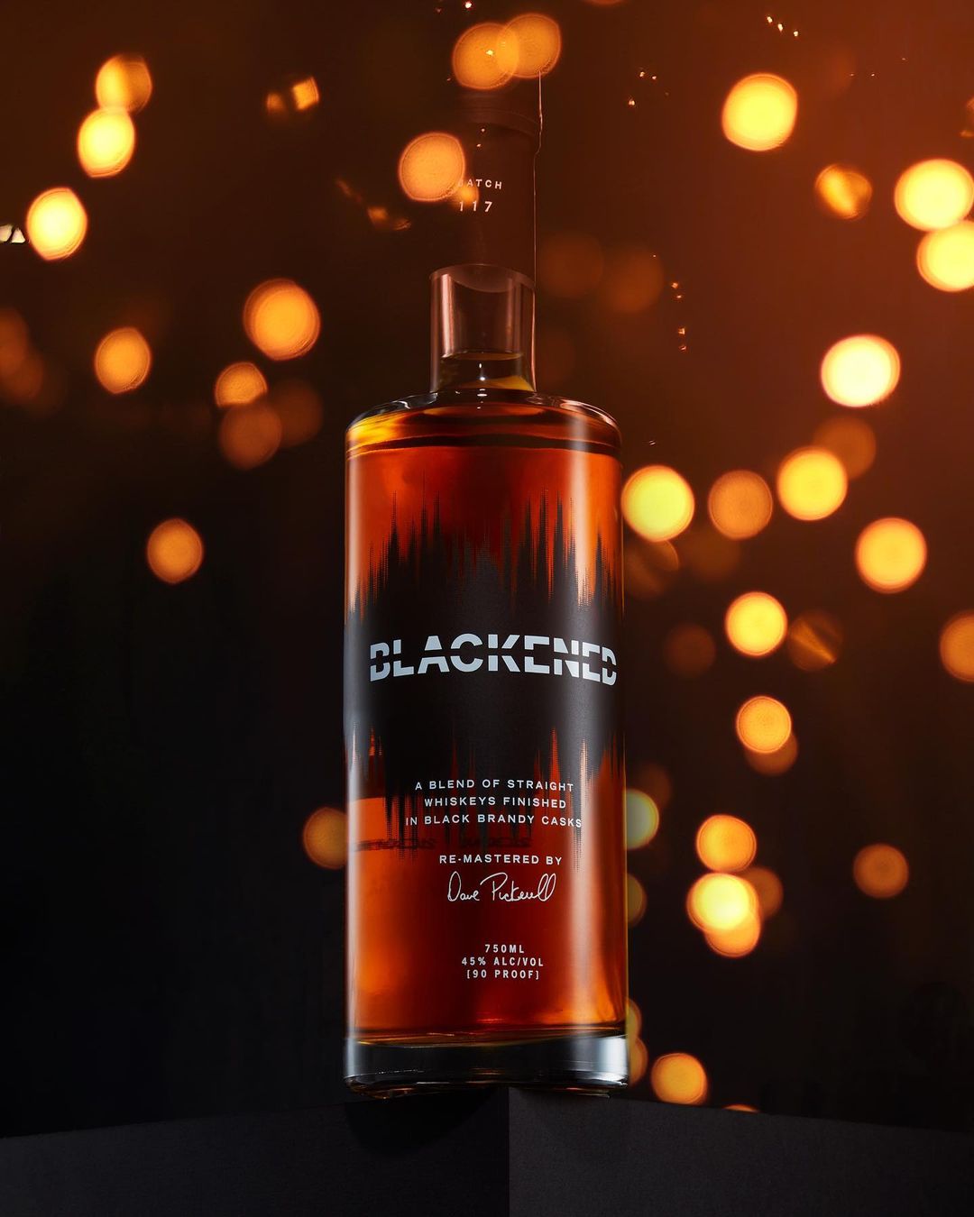 Angled shot of a Blackened Whiskey bottle on a dark surface, set against a backdrop of defocused golden bokeh lights.