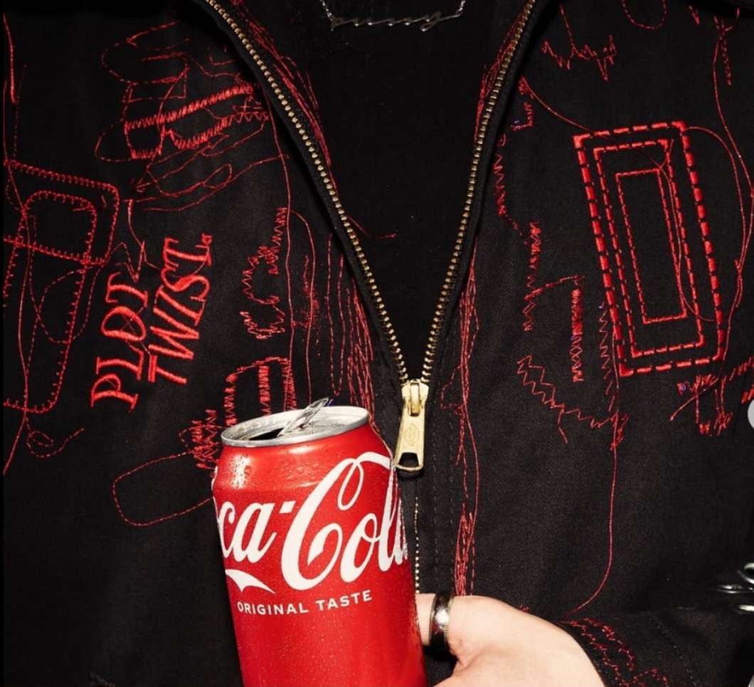 Close-up of Coca-Cola can held in hand against black jacket with red stitching details, showing brand logo and beverage context.