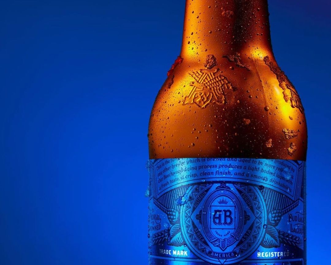 Close-up of a Budweiser beer bottle with condensation against a blue background. The bottle's label is visible, emphasizing the brand.