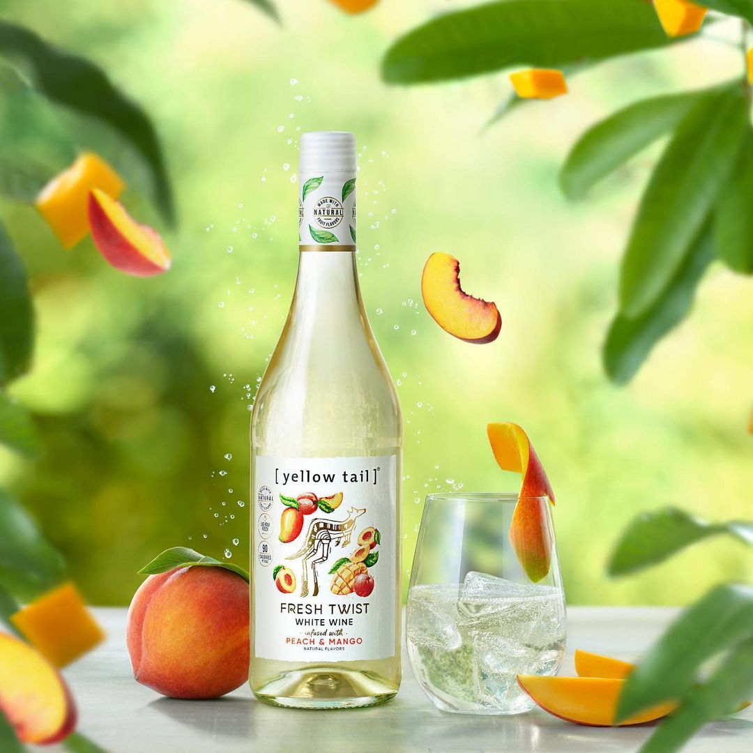 Bottle of Yellow Tail Fresh Twist white wine with peach and mango flavor, served with peach slices, ice, and green foliage background.