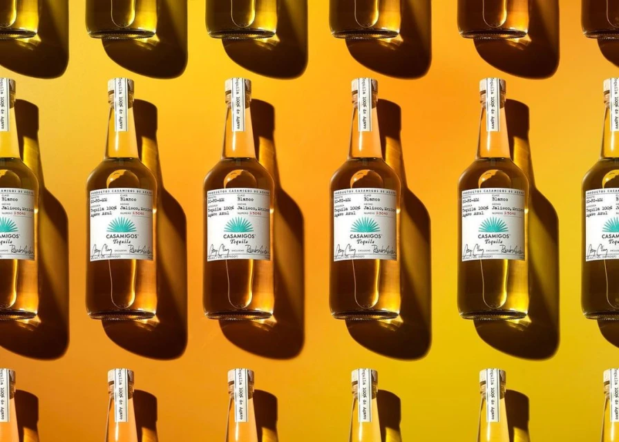 Multiple bottles of Casamigos Tequila sit in a repeating pattern against a yellow background, illuminated by shadows and bright lighting.