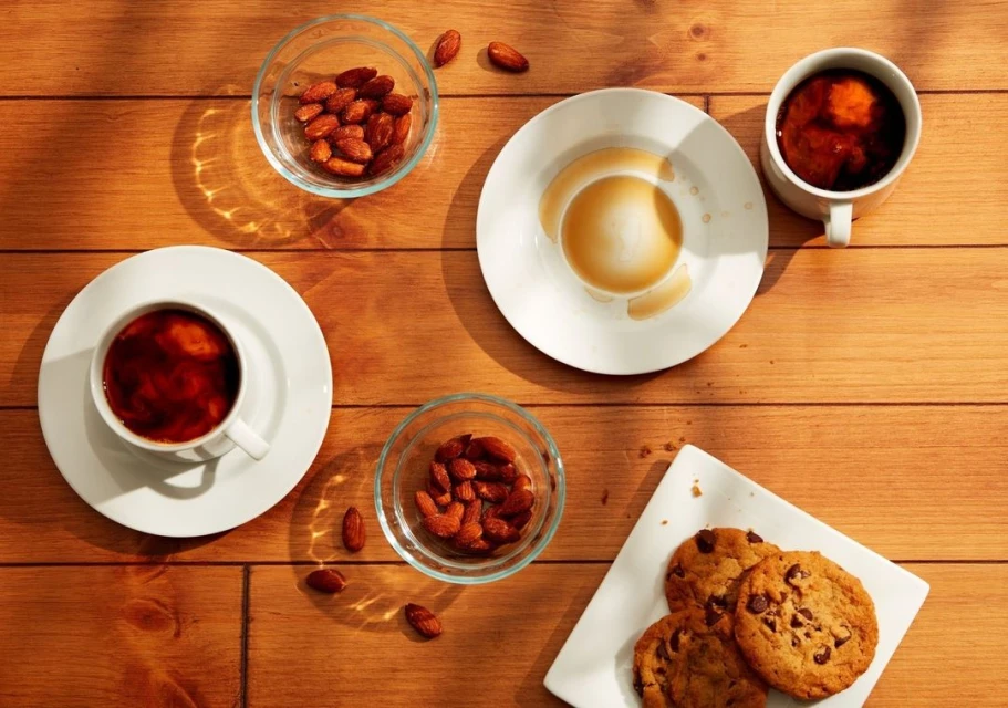 Overhead shot of wooden table featuring coffee cups, bowls of almonds, and chocolate chip cookies on a plate, warmly lit.