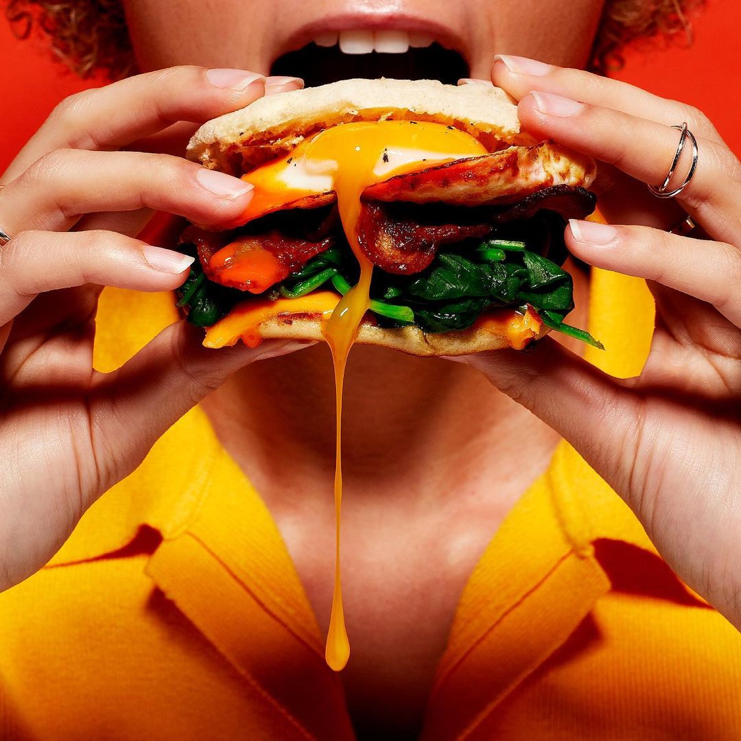 Close-up of a person holding a sandwich with a runny egg yolk, spinach, bacon, and cheese against a bright orange background.
