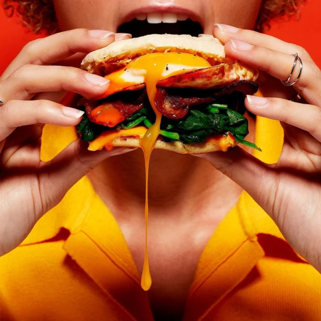 Close-up of a person holding a sandwich with a runny egg yolk, spinach, bacon, and cheese against a bright orange background.