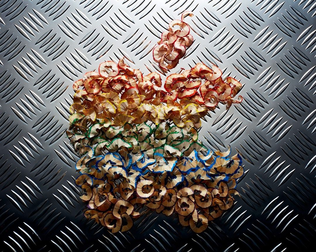 Apple logo formed from colored pencil shavings on a metallic diamond-plate surface, top-down composition, studio shot.