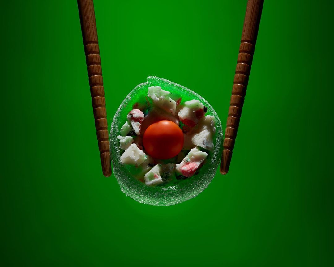 Overhead close-up of a sushi roll held between wooden chopsticks, featuring green seaweed, white rice, and a bright red center on green.