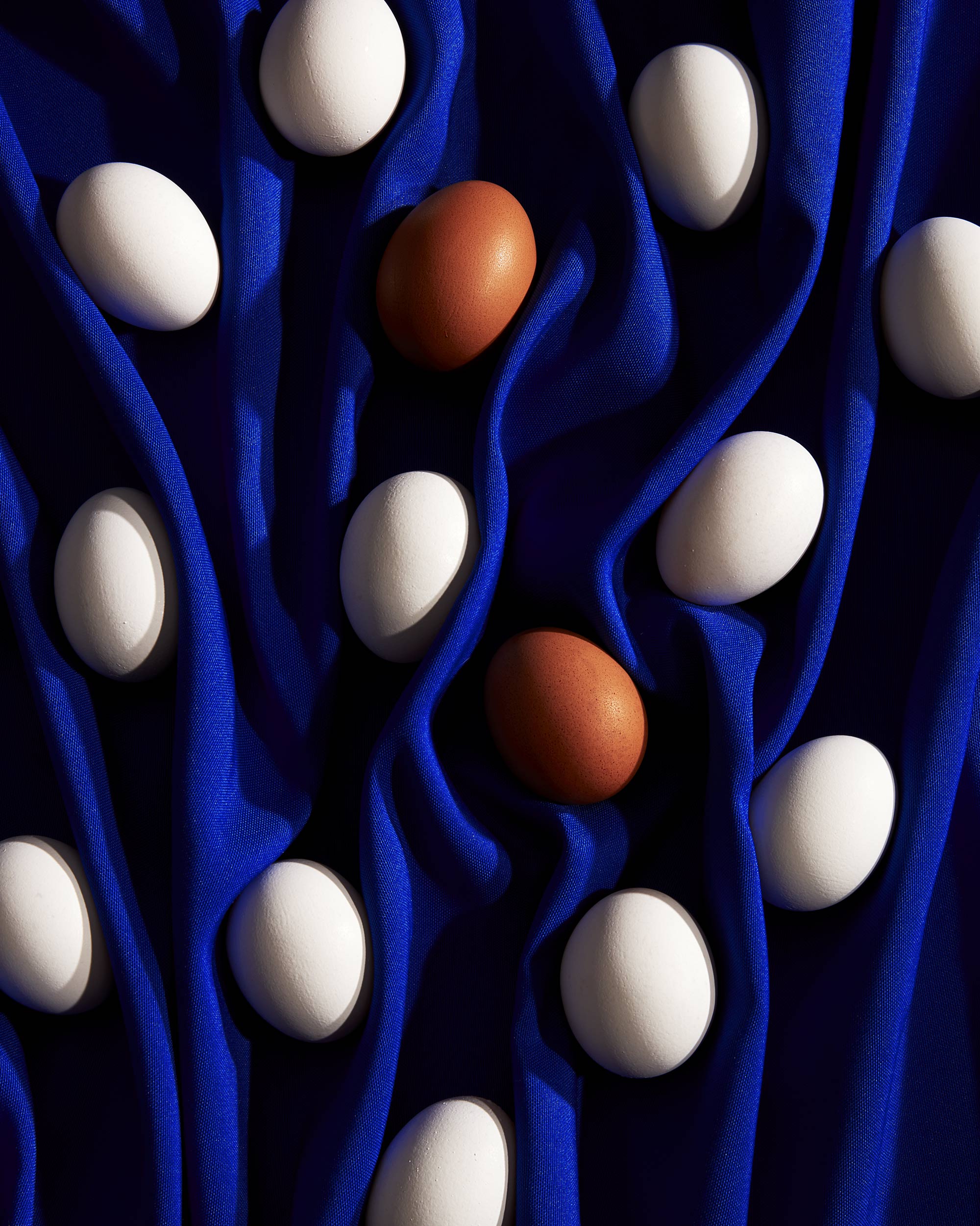 Still life studio shot of white and brown eggs arranged on a draped blue fabric, creating a textured and patterned surface.
