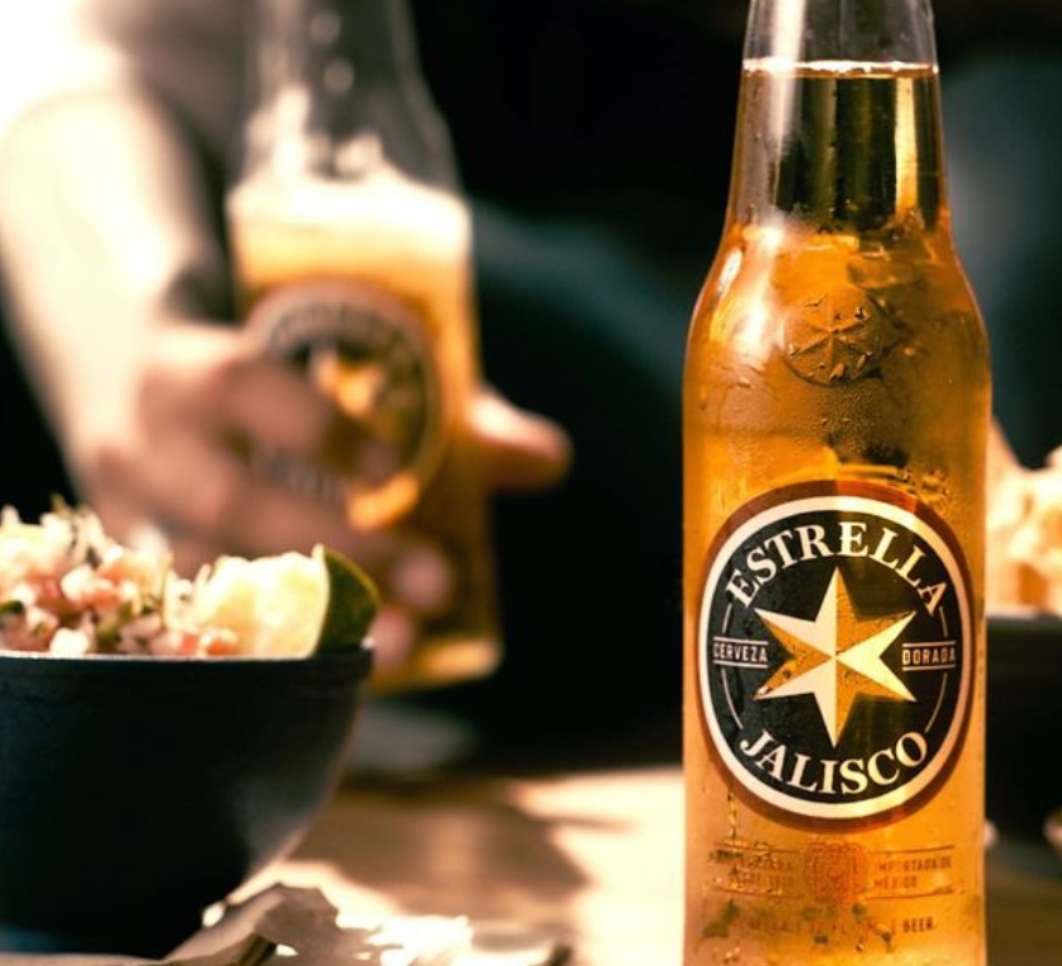 Close-up of a bottle of Estrella Jalisco beer with a bowl of ceviche and another beer in the background, creating an inviting food and beverage scene.