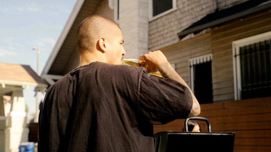 Man with a shaved head drinking from a bottle outdoors on a sunny day in a neighborhood, wearing a dark t-shirt with tattoo on arm.
