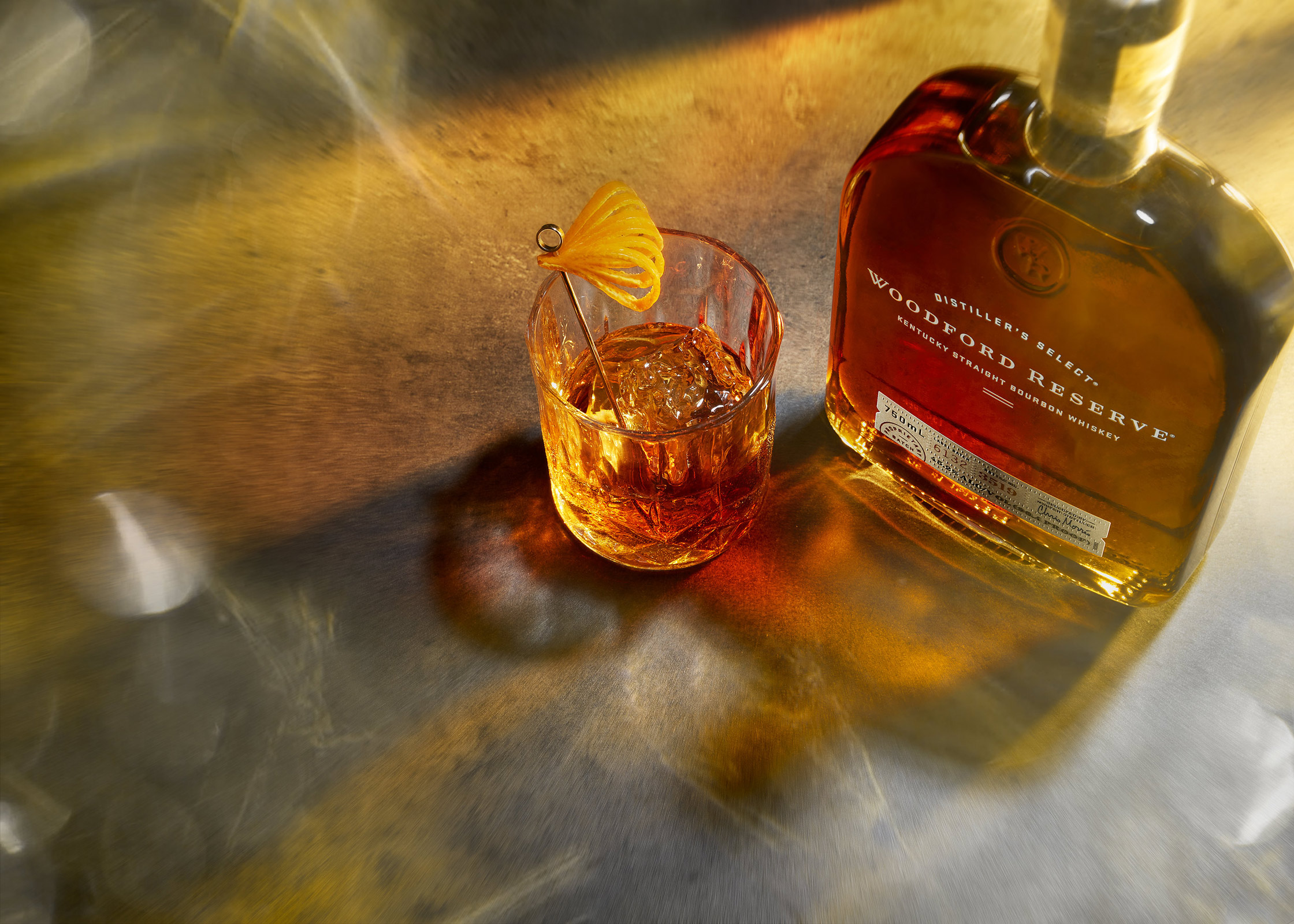 Close-up of a Woodford Reserve whiskey bottle next to a glass with ice and a lemon twist, set on a reflective surface.