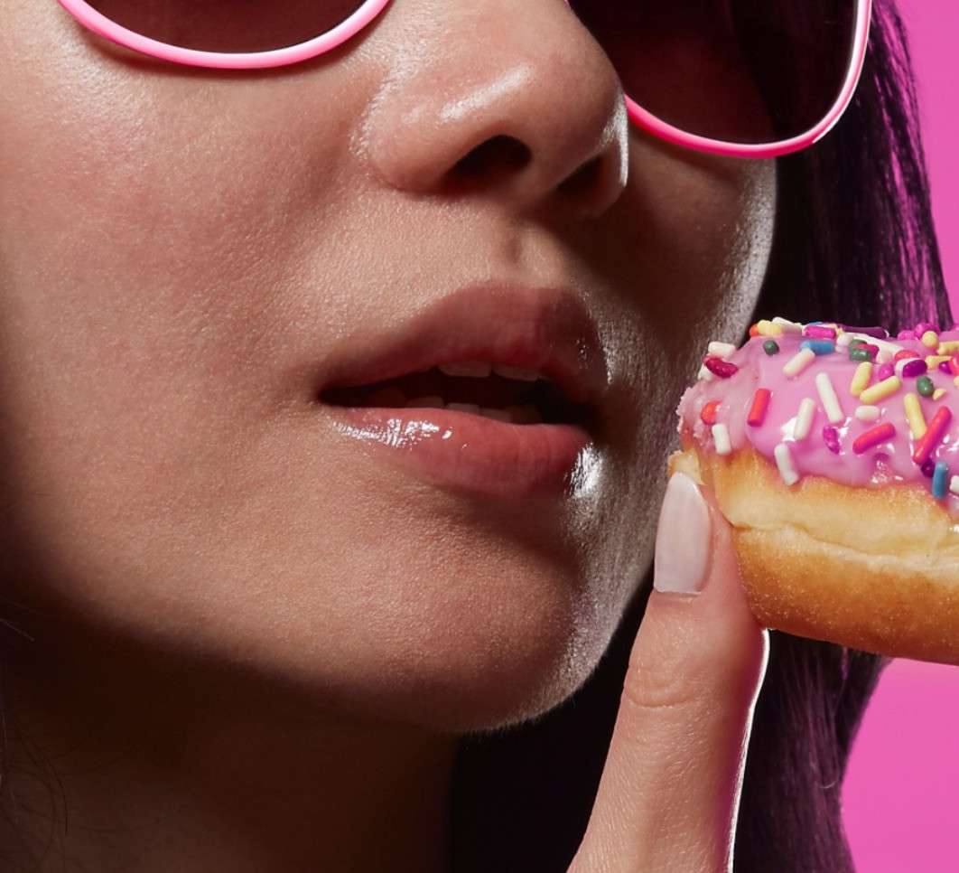 Close-up of a woman's face with pink sunglasses holding a doughnut with pink frosting and sprinkles against a pink background.