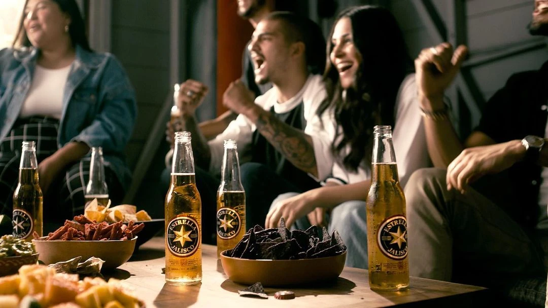 Enthusiastic group cheers at a table with Estrella Jalisco beer and snack bowls. Indoor setting, warm lighting, focus on shared celebration.